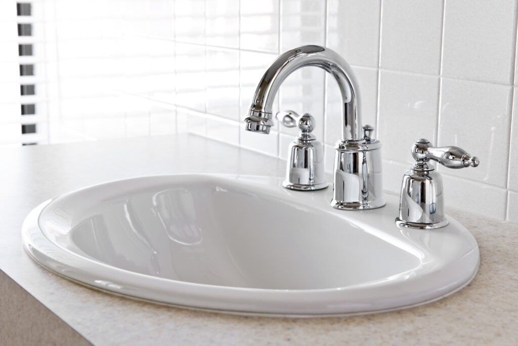 Newly installed white porcelain bathroom sink and chrome faucet. White tile wall behind counter.