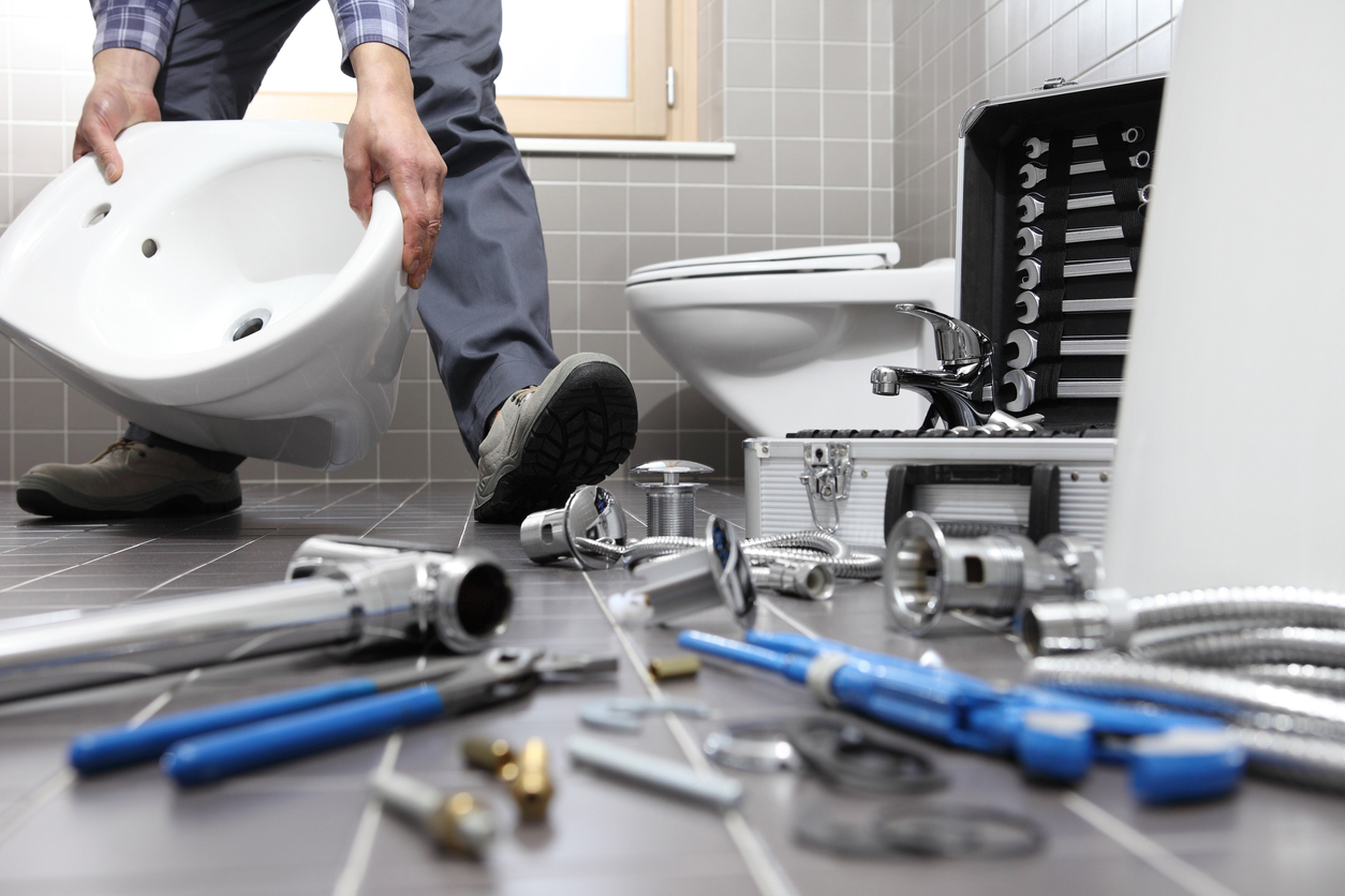 Cut view of a plumber installing a new toilet in a bathroom. Tools laid out across the gray tile floor.